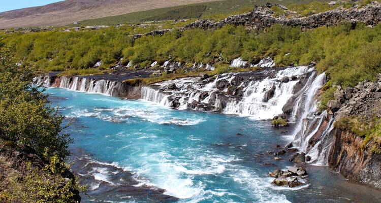 Trappende blauwe rivier die over lange vulkanische rotsrichels stroomt, omgeven door groen struikgewas.