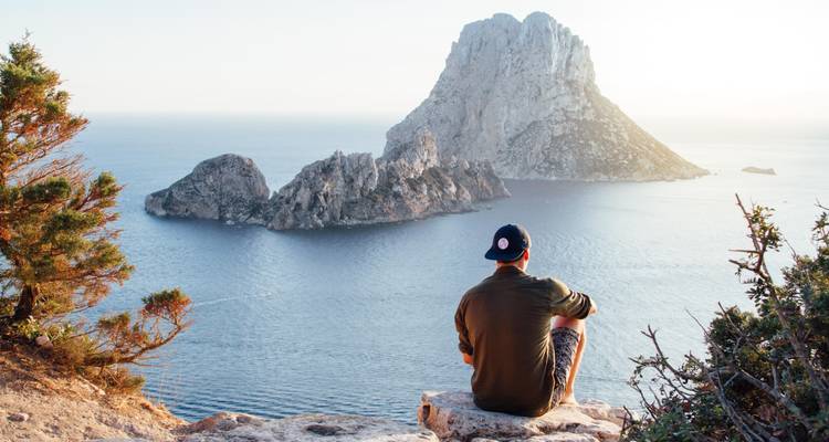 Un jeune homme assis au bord d'une falaise rocheuse contemple une île spectaculaire qui s'élève de la mer bleu profond.