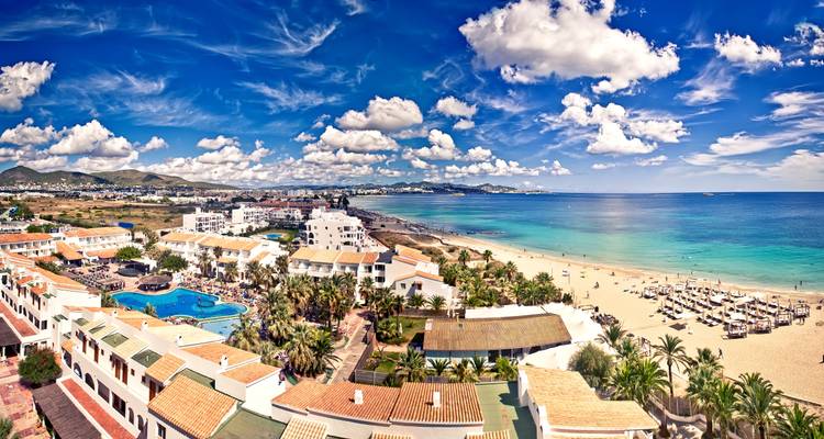 Station balnéaire côtière panoramique dotée de villas blanches, de piscines bordées de palmiers, d'une vaste plage de sable fin et d'une mer turquoise sous un ciel lumineux parsemé de nuages.