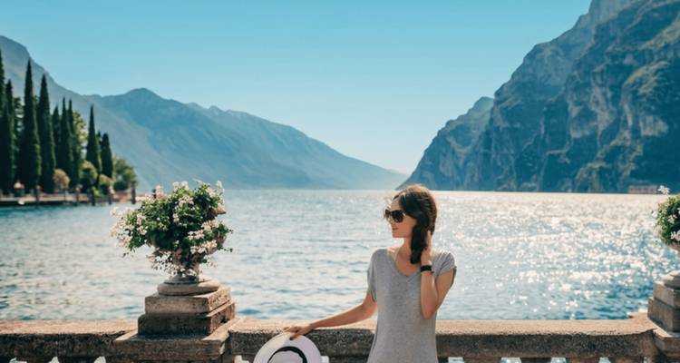Mujer con gafas de sol de pie junto a una barandilla con vistas a un tranquilo lago alpino rodeado de montañas escarpadas.