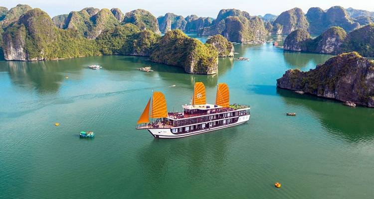 Un barco junco tradicional con velas naranjas se desliza por las aguas esmeraldas de la Bahía de Halong rodeado de islas de piedra caliza.