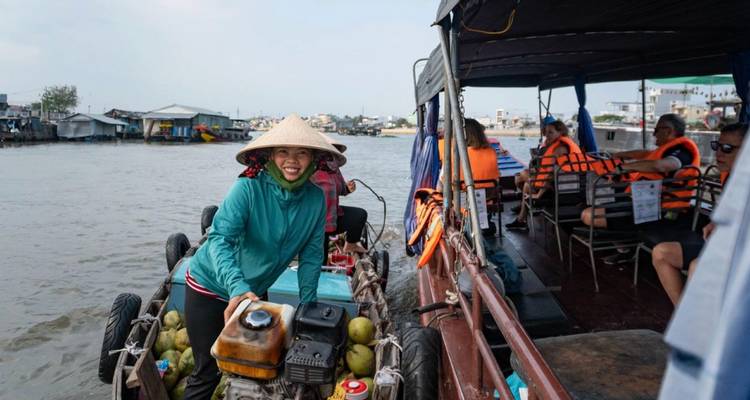 Un vendedor sonriente dirige una barca cargada de productos junto a turistas con chalecos salvavidas a bordo de un tour en bote por el Delta del Mekong.