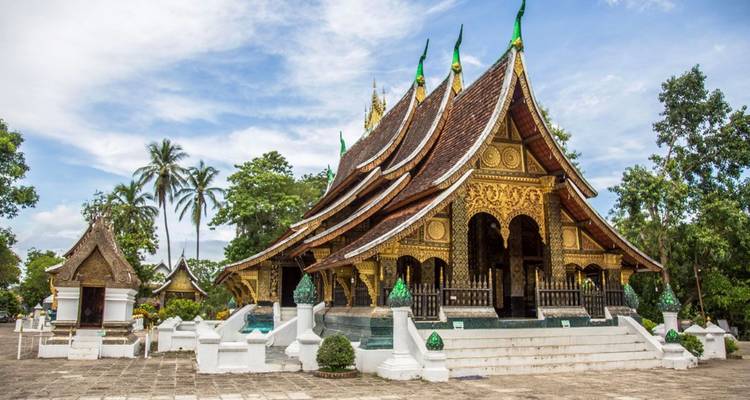 El techo dorado y ornamentado y los amplios aleros del Wat Xieng Thong se alzan entre la exuberante vegetación tropical bajo un cielo despejado.