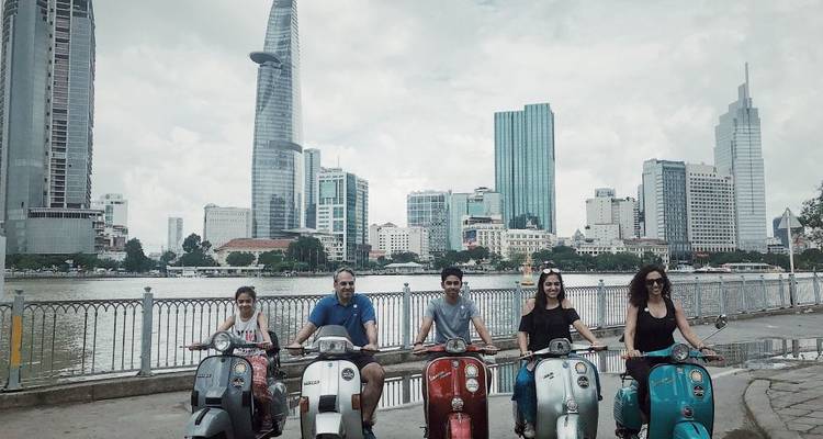 Fünf Reisende sitzen auf Vintage-Vespas entlang des Saigon-Flussufers mit Wolkenkratzern von Ho-Chi-Minh-Stadt im Hintergrund