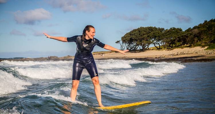 Surfeur en combinaison de plongée en équilibre sur une planche jaune chevauchant une petite vague près d'une rive sablonneuse