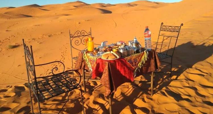 Table de petit-déjeuner dressée avec du thé marocain, des pâtisseries et du jus posés sur les dunes de sable doré du Sahara