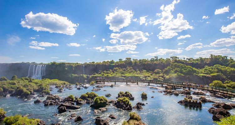 Weitläufige Aussicht auf die Iguazu-Wasserfälle mit Nebel, üppigem Dschungel und einer Aussichtsbrücke über dem Fluss.