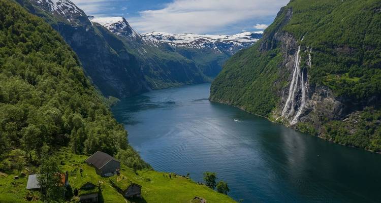 Un fjord bleu émeraude sinuant entre des montagnes escarpées et boisées avec une haute cascade se déversant le long de la paroi rocheuse.