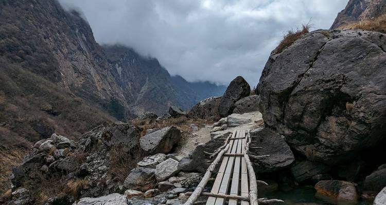 Un estrecho sendero de tablones de madera serpentea entre rocas hacia un desfiladero montañoso envuelto en niebla.