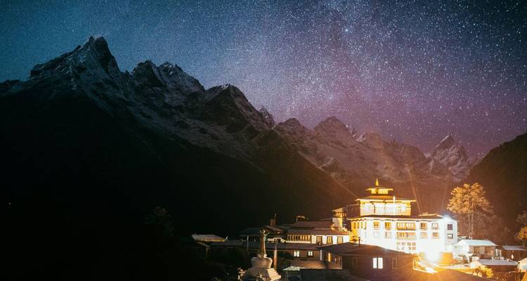 Un monasterio himalayo de noche brilla calurosamente bajo un cielo lleno de estrellas y picos dentados y oscuros.