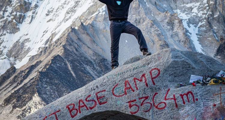 Un trekker triunfante se para en la cima de una roca tallada marcada con 'EBC 5364m' con una pared helada del Himalaya de fondo.