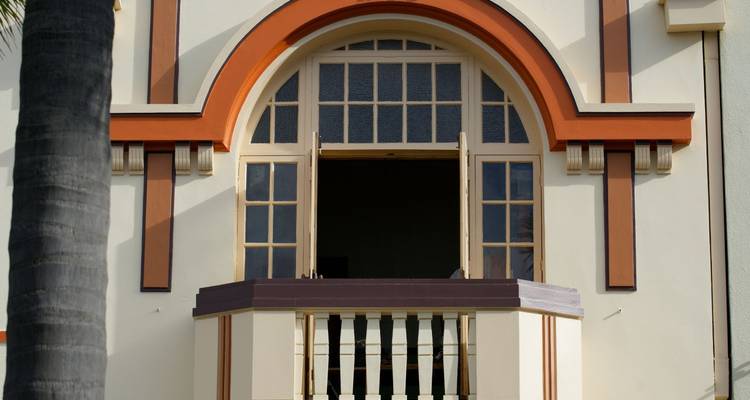 Detail van een room- en terracottakleurige Art Deco-balkon en boogvormige ramen met palmboomstam aan de zijkant.