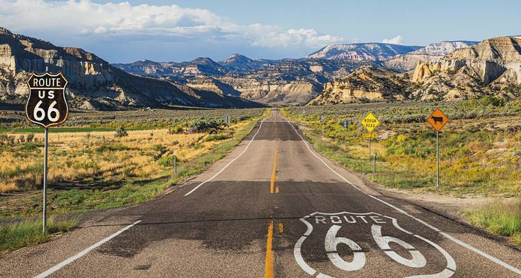 Carretera vacía de la Ruta 66 dirigiéndose hacia montañas desérticas escarpadas con señales clásicas de carretera pintadas en el asfalto.