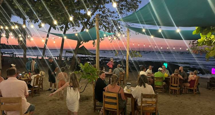 Restaurante de playa al aire libre al atardecer con luces de cuerda y comensales sentados en la arena