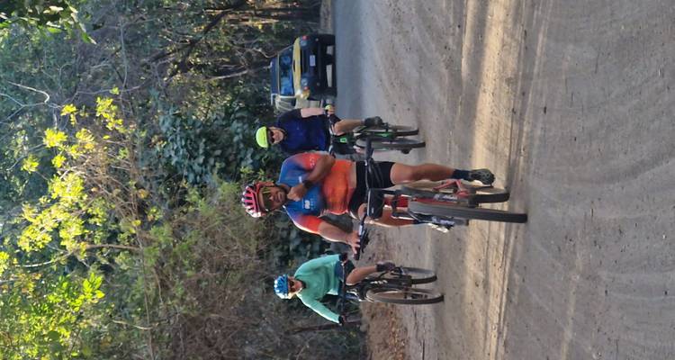Ciclistas andando en un camino de tierra sombreado por bosque seco con vehículo de apoyo detrás