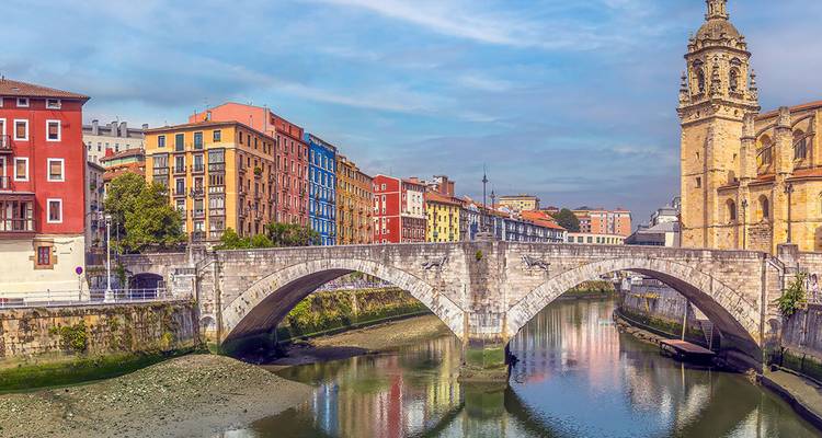 Un pont en arche de pierre enjambe une rivière reflétante flanquée de bâtiments historiques colorés et d'un clocher d'église à Bilbao.