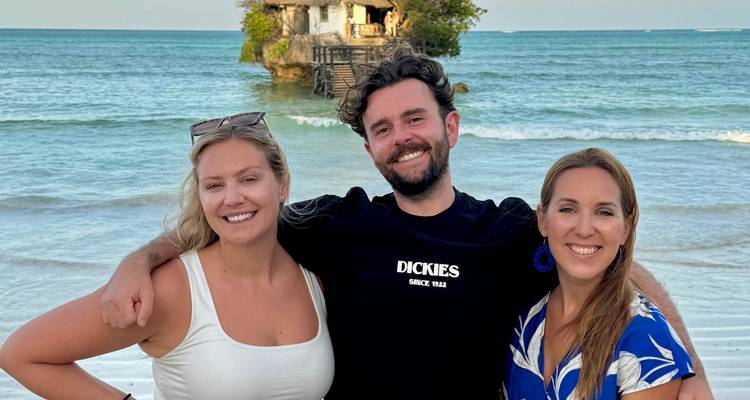 Tres amigos sonriendo en una playa con el restaurante The Rock sentado en un pequeño islote frente a la costa detrás de ellos