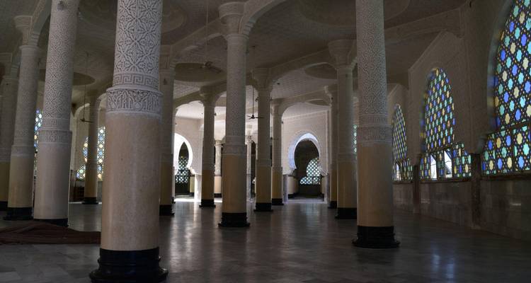 Hall intérieur faiblement éclairé d'une mosquée avec de hautes colonnes décorées et des fenêtres cintrées en vitrail.