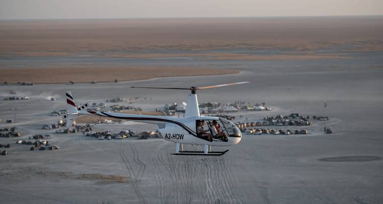 Small helicopter soars above vast grey salt pan dotted with expedition vehicles at dusk.