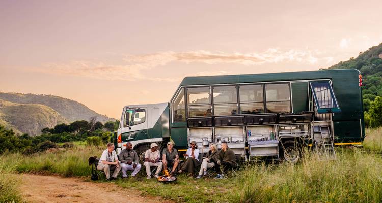 Travellers relax around campfire beside overland truck amid green hills at sunset.