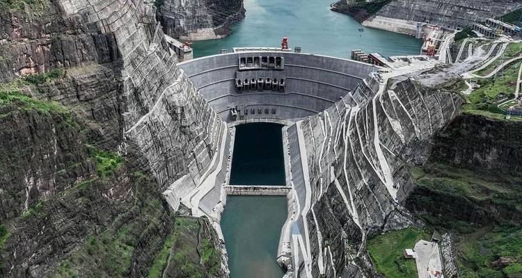 Aerial view of a massive concrete dam stretching between steep canyon walls with turquoise water on both sides.