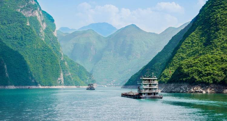 Cargo boats glide along a jade-green river flanked by towering misty mountains in a dramatic gorge.