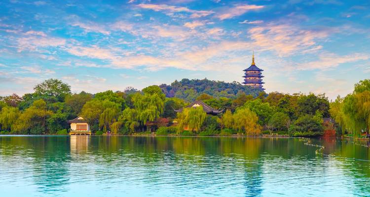 Serene lake bordered by lush trees with a hilltop pagoda rising above under a colorful sky at golden hour.