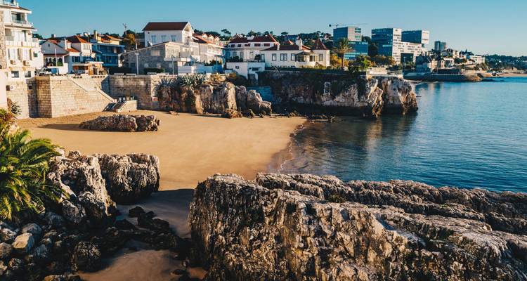 Tranquila cala de arena bordeada por afloramientos rocosos y un pueblo costero reflejándose en la luz tranquila de la mañana.