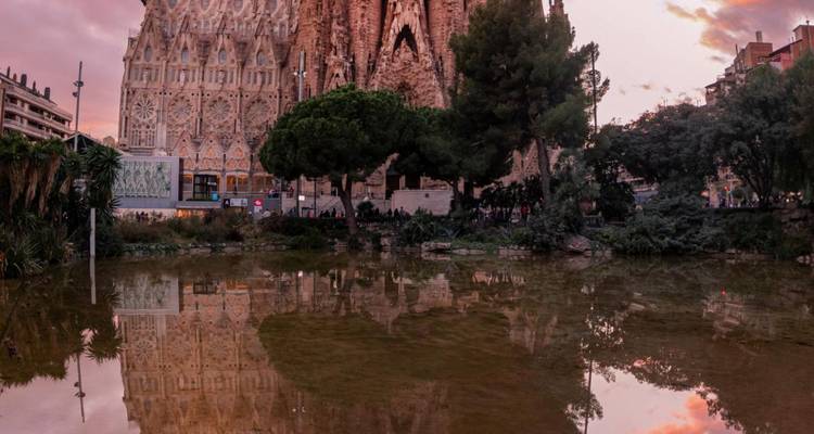 Vue en soirée de la Sagrada Família reflétée dans un bassin sous un ciel de coucher de soleil rose et violet.