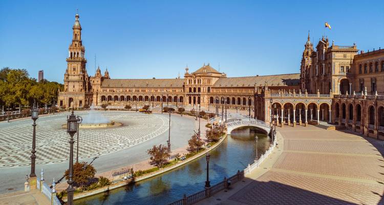 Vue panoramique large de la Plaza de España avec ses bâtiments ornés, son pont en arche et son canal sous un ciel bleu dégagé.