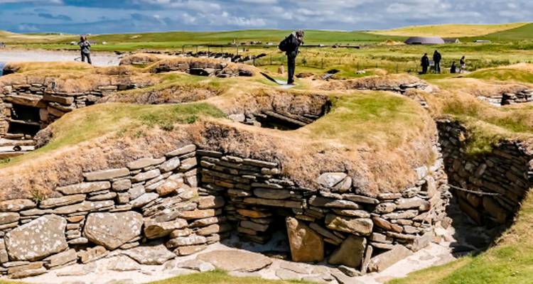Grass-covered stone dwellings of the Neolithic village Skara Brae with visitors exploring.