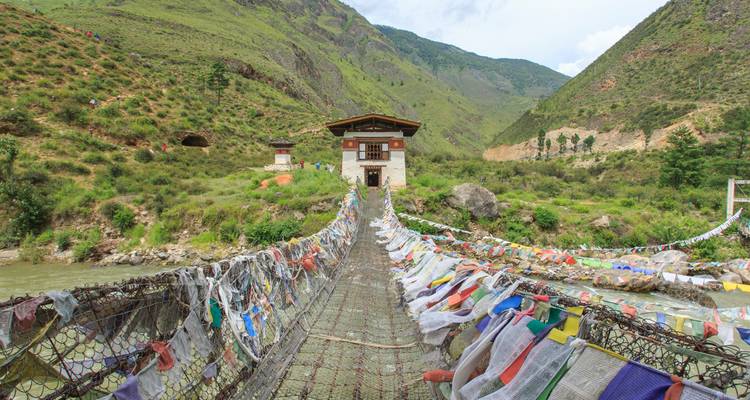 Puente colgante decorado con banderas de oración multicolores que lleva a una pequeña casa bhutanesa en un valle exuberante.