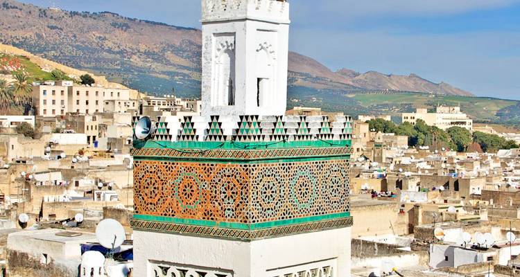 Minarete decorativo de azulejos verdes que se eleva sobre los tejados de Fes con montañas al fondo.