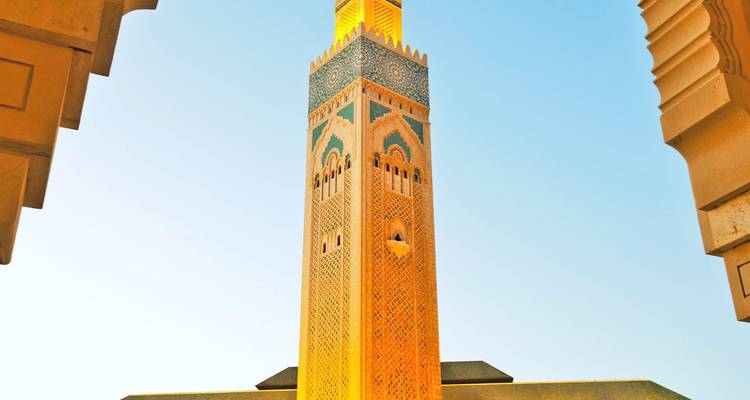 Vista hacia arriba del minarete iluminado de la Mezquita Hassan II enmarcado por arcos de arenisca contra un cielo despejado.