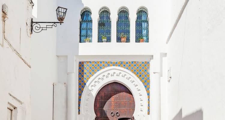 Puerta marroquí encalada enmarcada por coloridos azulejos zellige y una puerta de madera con arco bajo ventanas arqueadas.
