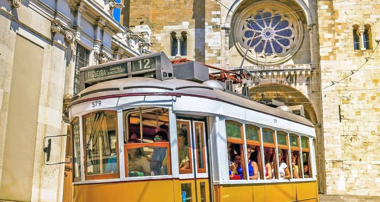Klassieke gele tram vol passagiers die langs de rozetvenster-gevel van de Kathedraal van Lissabon rijdt.
