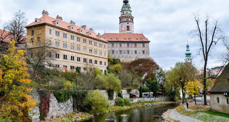 Vue de la rivière d'un complexe de château historique tchèque avec des arbres d'automne se reflétant dans l'eau sous un ciel couvert.