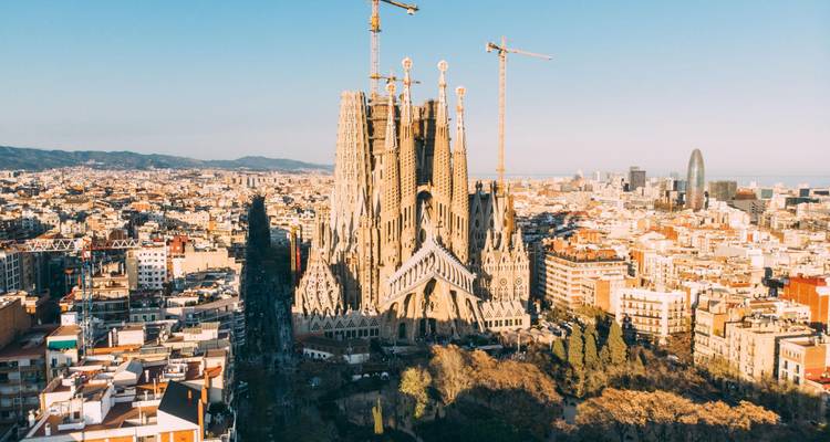 Vista aérea de Barcelona dominada por la imponente Sagrada Familia con bloques de ciudad extendiéndose hacia las montañas lejanas.