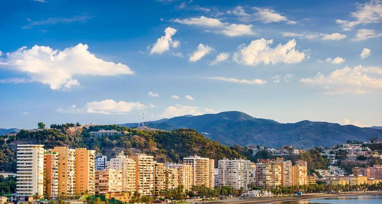 Skyline costero de Málaga con bloques de apartamentos modernos respaldados por colinas verdes y nubes dispersas en el cielo azul.