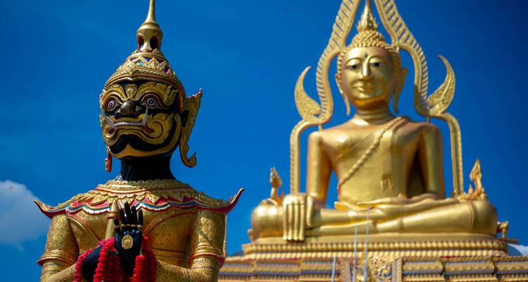 Nahaufnahme einer farbenfrohen Schutzdemonfigur neben einer großen sitzenden goldenen Buddha-Statue vor lebendigem blauem Himmel in Thailand.