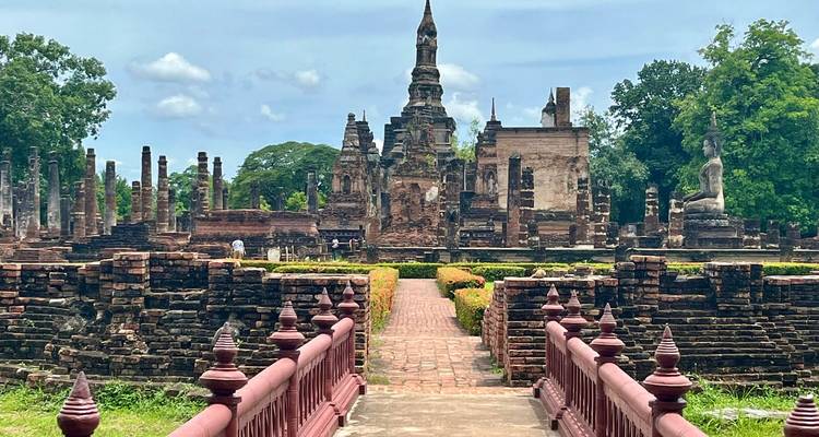 Zerstörte Backsteinsteupas und Säulen des antiken Historischen Parks von Sukhothai, betrachtet von einer roten Brücke über dem Wassergraben.