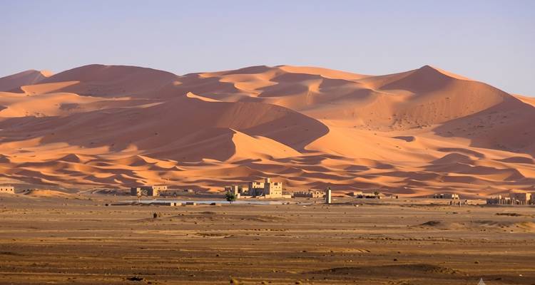 Sweeping golden sand dunes of the Sahara with a small desert settlement at their base near Merzouga.