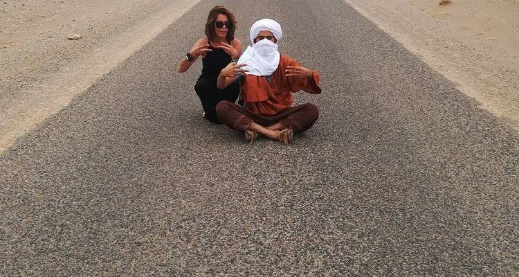 Two travelers sit cross-legged on an empty desert highway making playful hand gestures.