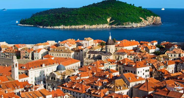 Vista cercana de los tejados anaranjados de Dubrovnik con cúpula y torres de iglesia recortadas contra el mar azul profundo y la isla.