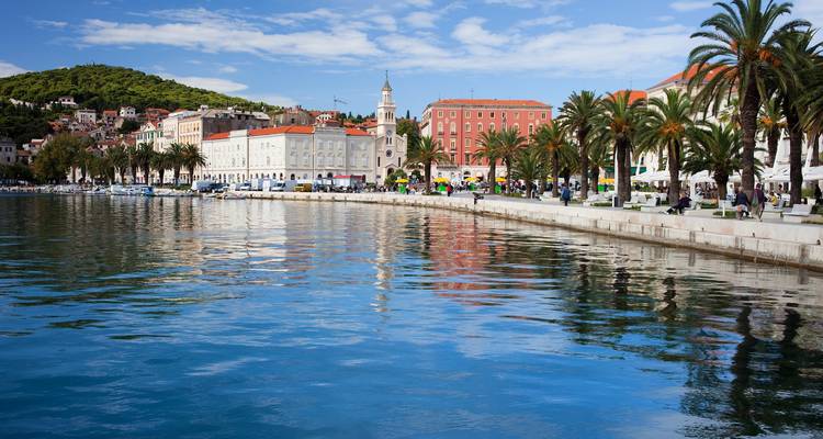 Paseo junto al mar bordeado de palmeras y edificios coloridos se reflejan en las aguas tranquilas del puerto histórico de Split.