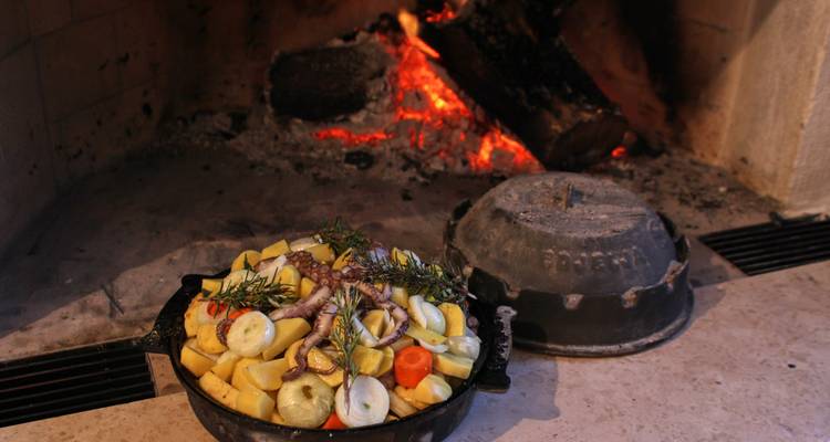 Traditional Croatian peka dish with vegetables and meat cooks in a cast-iron pot beside an open hearth.