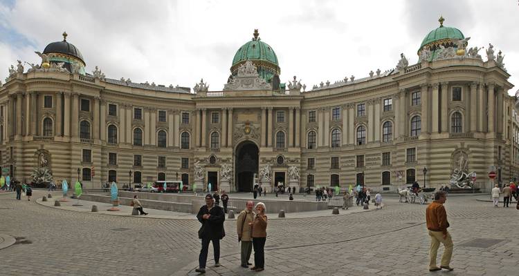 Amplia fachada del Palacio de Hofburg de Viena con turistas de pie en la plaza empedrada.