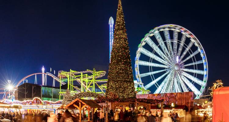 Escena nocturna festiva de una feria navideña con un árbol iluminado alto, montaña rusa y una rueda de la fortuna gigante.