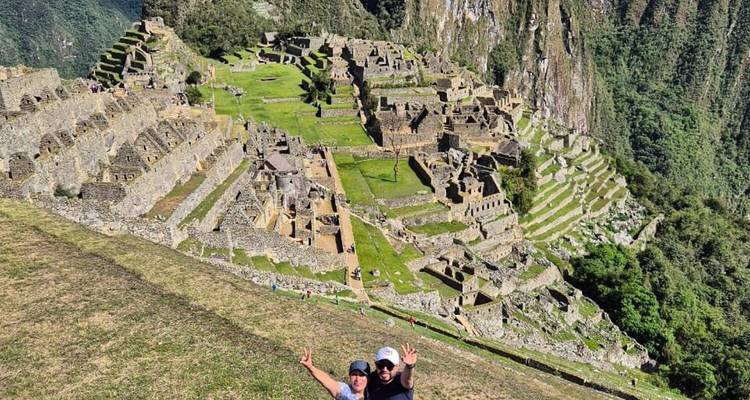 Un couple fait un geste de la main vers la caméra avec une vue panoramique des terrasses et des pics de Machu Picchu en arrière-plan.