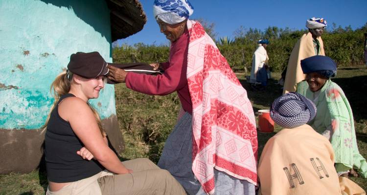 Mujeres locales comparten comida con una viajera fuera de una rondavel turquesa bajo cielos despejados.
